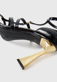 Sandal de salto alto em pele sintética preta com tiras finas e um salto metálico dourado em forma de trompete alargado, com uma sola texturizada.