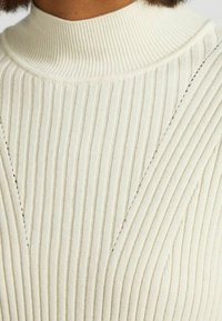 Krämmig ribbad stickad tröja med hög krage och diagonala stickningar som ger struktur och textur till designen.