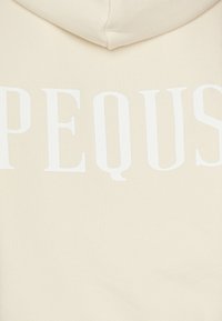 Krema barve kapuca s mehkimi, teksturiranimi vlakni. Opremljena je z drzno belim napisom "PEQU". Vključuje kapuco za dodatno zaščito.