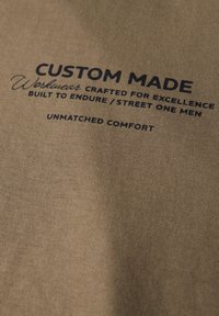 Béžová látka s hladkým texturem, na které je vytištěn text: "CUSTOM MADE Pracovní oděvy vyrobené pro excelenci, navržené pro odolnost / Street One Muži BEZKONKURENČNÍ POHODLÍ."