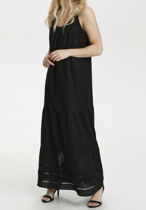 Kobieta w czarnej, bezrękawnej sukni maxi z warstwowym dołem i czarnych sandałach na wysokim obcasie, stojąca na tle jednolitego białego tła.