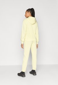 Κίτρινο φούτερ με κουκούλα και ταιριαστό παντελόνι φόρμας από μαλακό ύφασμα. Διαθέτει κουκούλα, ριμπ μανσέτες και άνετη εφαρμογή. Μαύρα παπούτσια ολοκληρώνουν το ντύσιμο.