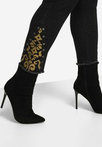 Los jeans de mezclilla negros presentan acentos dorados en la parte inferior de la pierna, adornados con tachuelas plateadas. Botas de ante negras de tacón alto con punteras puntiagudas.