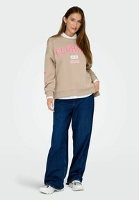 Sweatshirt beige avec "FLORIDA MIAMI EST. 1945" en lettres roses, associé à un jean évasé bleu et des baskets blanches. Texture lisse, coupe décontractée.
