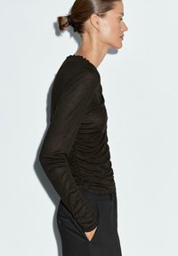 Maglietta a maniche lunghe di colore marrone scuro con design aderente, caratterizzata da arricciature laterali e scollatura ondulata, abbinata a pantaloni neri.