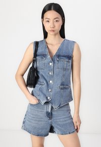 Denimli yelek, V yaka, kolsuz tasarım ve ön düğmelerle. Taş yıkama açık mavi renkte, iki göğüs cebi ve kavisli etek ucu ile.