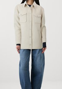 Chaqueta de camisa de mezcla de lana beige con cuello, botones frontales, dos bolsillos en el pecho y un ajuste relajado, combinada con jeans de pierna ancha de color azul claro.