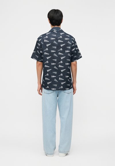 Άνδρας που φοράει κοντομάνικο σκούρο πουκάμισο με λευκά τρίγωνα και ανοιχτό μπλε τζιν, στέκεται με την πλάτη γυρισμένη σε απλό φόντο.