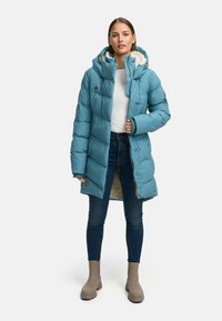 Turkusowa kurtka typu puchówka z miękkim, polarowym kapturem, napami oraz długością do kolan. Połączona z ciemnymi dżinsami i beżowymi botkami za kostkę.