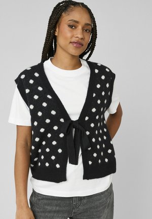 Jeune femme aux cheveux tressés portant un gilet noir en tricot à pois blancs sur un t-shirt blanc et un jean gris, debout devant un fond uni.
