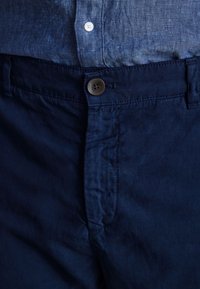 Pantaloni chino de culoare bleumarin, cu o textură netedă, având un sistem de închidere cu buton și detalii de cusătură subtile de-a lungul taliei și buzunarelor.