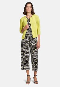 Żółta lniana kurtka na czarno-białym kombinezonie w wzory z żółtymi akcentami. Kombinezon ma szerokie nogawki i krótkie rękawy. Czarne szpilki.