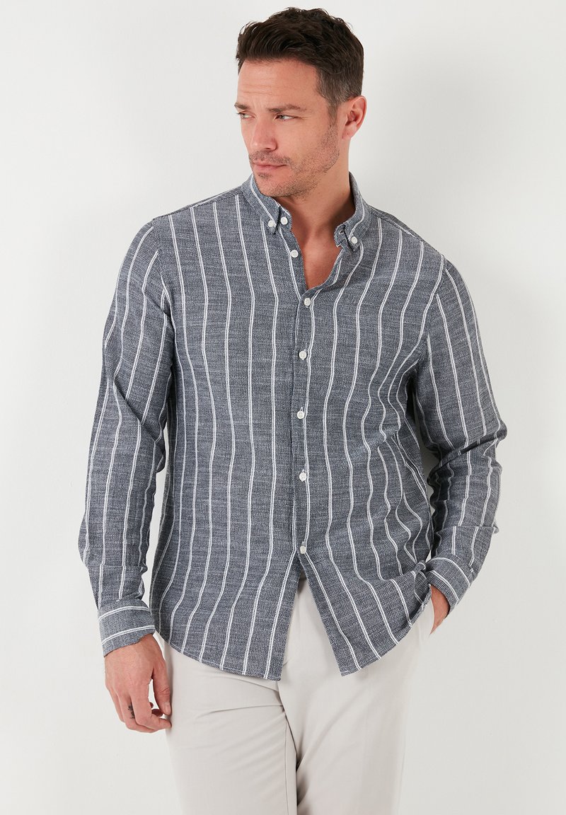 Camisa de botón a rayas azul y blanca con cuello, mangas largas y tejido texturizado. Estilizada con pantalones de color claro.