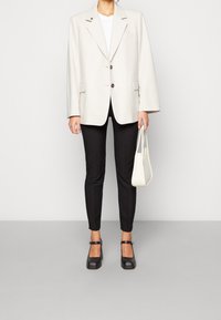 Blazer beige oversize con due bottoni, pantaloni neri attillati, scarpe Mary Jane nere e una piccola borsa bianca. Texture liscia in tutto.