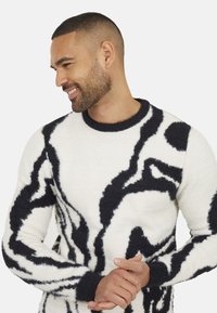 Pull crème avec de bold motifs abstraits noirs, un col rond et un tissu texturé. Présente des poignets et un ourlet sombres en couleur contrastante.