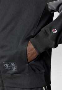 Μαύρο αθλητικό sweatshirt με γκρι λεπτομέρεια, που διαθέτει λογότυπο και μια καφέ εσωτερική τσέπη. Οι ριμπ μανσέτες και το στρίφωμα παρέχου�ν δομή.