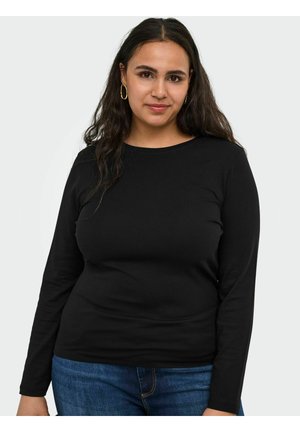 Vrouw met lang donker haar, gouden oorringen, een zwart shirt met lange mouwen en een blauwe jeans, staand tegen een effen lichte achtergrond.