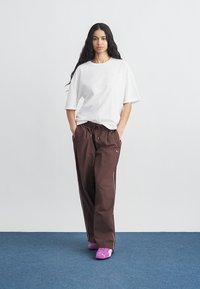 Γυναίκα με μακριά σκούρα μαλλιά που φορά λευκό υπερμεγέθες T-shirt, καφέ φόρμα και ροζ αθλητικά παπούτσια Puma, στέκεται σε μπλε χαλί.