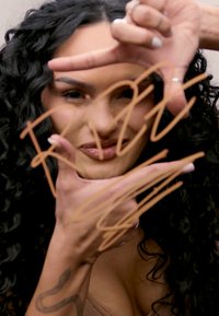 Una persona con i capelli ricci posa con le mani che incorniciano il volto. Un motivo astratto è disegnato su una superficie di un caldo tono marrone.