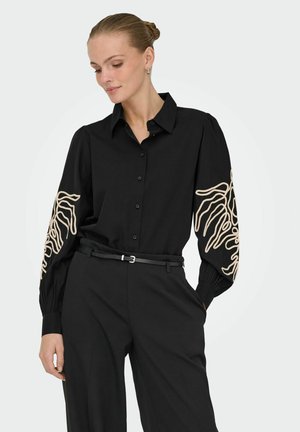 Femme portant un chemisier noir à boutons avec une broderie abstraite beige sur les manches bouffantes, un pantalon noir taille haute et une ceinture noire mince.