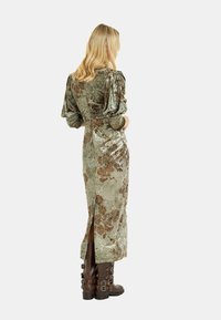 Langes, grünes Samtkleid mit Puffärmeln und floralem Muster in Bronze. Verfügt über einen Schlitz an der Seite und wird mit braunen, mit Schnallen verzierten Stiefeln kombiniert.