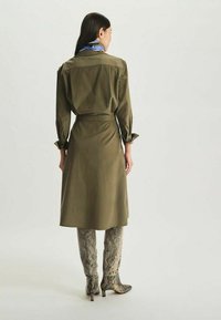 Robe chemise vert olive à manches longues et poignets noués, avec une taille cintrée et une jupe fluide. Associée à des bottes montantes à imprimé serpent beige.