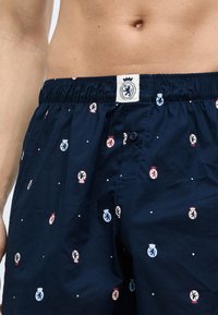 Mørkeblå shorts lavet af letvægtsstof med en knap. Har et gentaget våbenskjoldmønster i rød og lyseblå på en prikket baggrund.