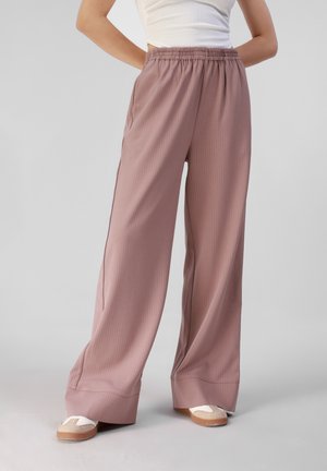 Persona con pantalones anchos de color rosa polvoriento con cintura elástica y zapatillas blancas, de pie frente a un fondo claro y liso.