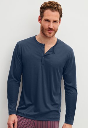 Man met kort krullend haar en baard draagt een donkerblauw henley shirt met lange mouwen en rode gestreepte broek tegen een effen lichte achtergrond.