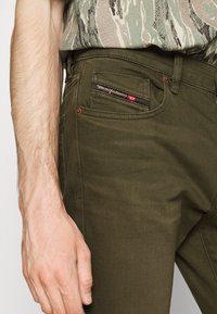 Pantalones de mezclilla verde oliva con corte ajustado, que presentan una etiqueta bordada en el bolsillo trasero en rojo y negro, y remaches de cobre en los bolsillos.