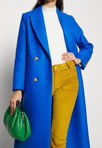 Cappotto doppiopetto blu brillante, dolcevita bianco a coste, pantaloni in corduroy dorati e una borsa verde arricciata con una chiusura metallica.