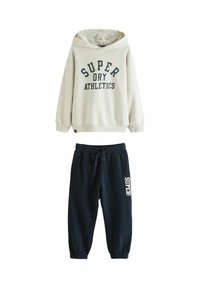 Bežs kapuces džemperis ar tekstu "SUPER DRY ATHLETICS" kopā ar melnām sporta biksēm, kurām ir sānu kabatas un teksts "SUPER" uz kājas.