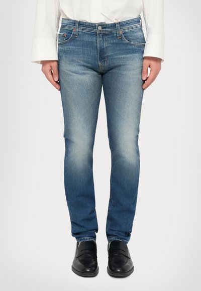 Hellblaue Jeans aus Denim mit schlanker Passform, verblasstem Textur, Fünf-Taschen-Design und Kontrastnähten, kombiniert mit schwarzen Schuhen.