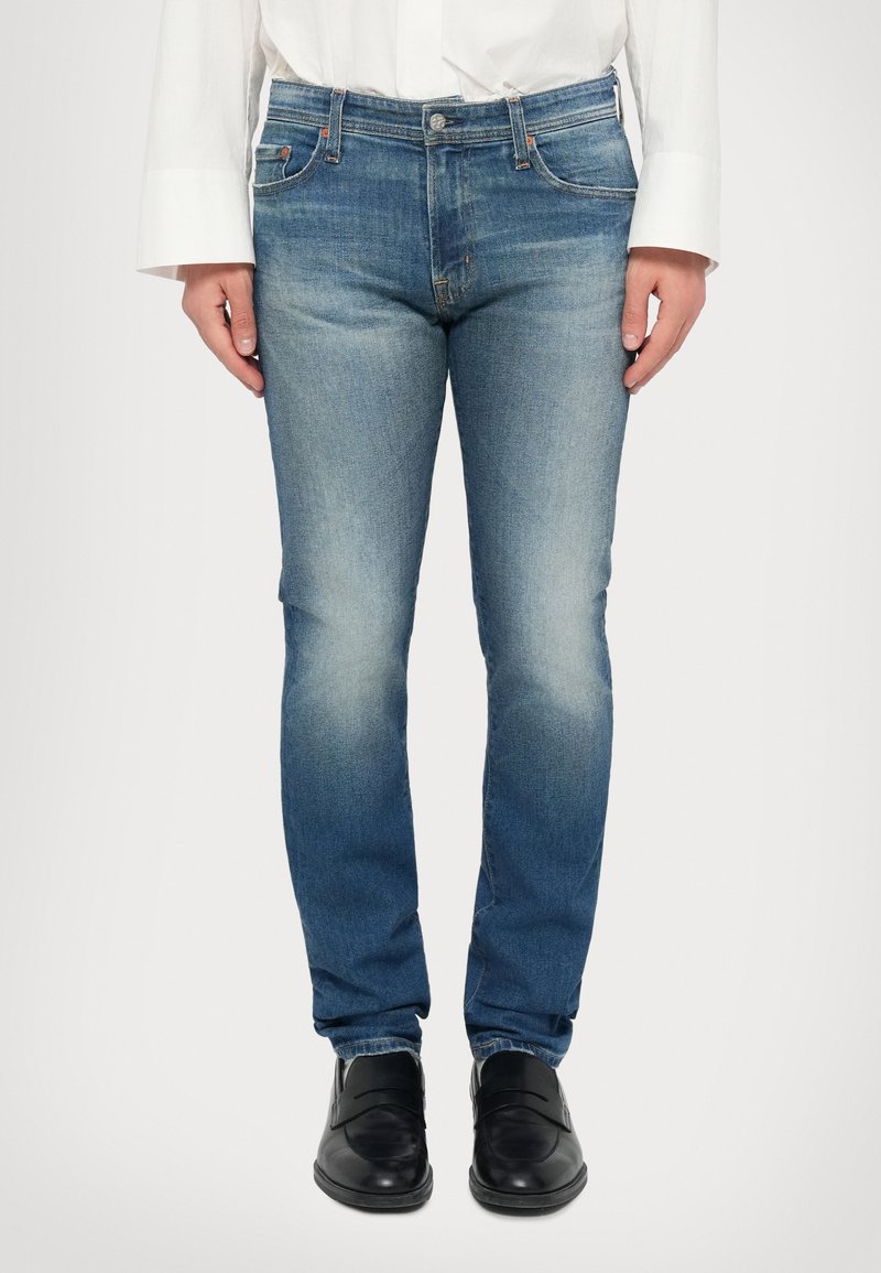 Světlé modré džíny s slim fit střihem, vybledlou textura, designem s pěti kapsami a kontrastním šitím, v kombinaci s černými botami.