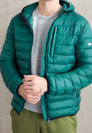 Person trägt eine petrolfarbene gesteppte Jacke über einem grauen Hemd, olivgrüne Hose, die Hände sind verschränkt und zeigen ein "TIME"-Tattoo am Finger.