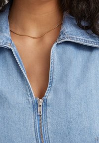 Ljusblå denimjacka med dragkedja fram och krage. Anmärkningsvärd textur med synliga sömmar och ett enkelt halsband i guldkedja.