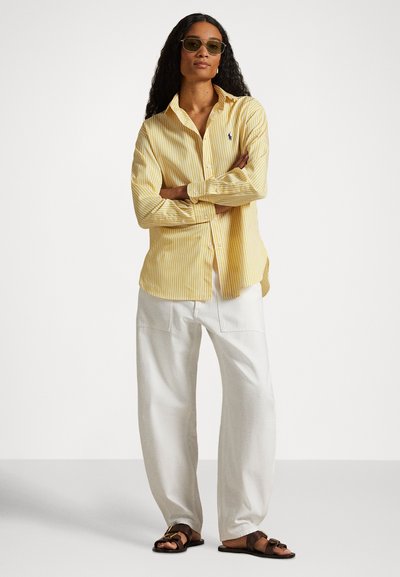 Sieviete ar gariem cirtainiem matiem, valkājot zaļganus saulesbrilles, dzeltenu svītrainu kreklu, baltas bikses un brūnas sandales, stāv ar sakrustotām rokām.