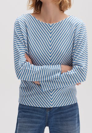 Femme aux cheveux blonds courts portant un pull rayé blanc et bleu et un jean bleu, bras croisés au niveau de la taille, tête tournée sur le côté.