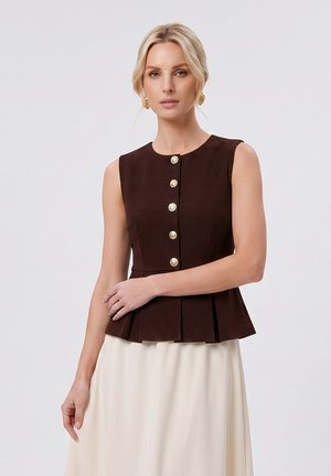 Blond vrouw met een mouwloze donkerbruine top met knopen, gouden oorbellen en een lichtbeige rok, poserend tegen een effen achtergrond.