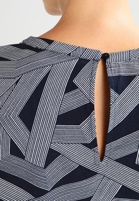 Blusa blu navy con un motivo geometrico a strisce bianche, dettaglio a chiave sulla schiena e classico scollo tondo. Tessuto morbido.