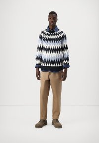 Maglione lavorato a maglia con motivo a zigzag in nero, bianco e blu, indossato sopra una camicia con colletto in denim, abbinato a pantaloni beige e stivali marroni.