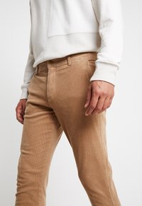 Beżowe spodnie sztruksowe o wąskim kroju, z przodem zapinanym na guziki i bocznymi kieszeniami, zestawione z jasną bluzą.