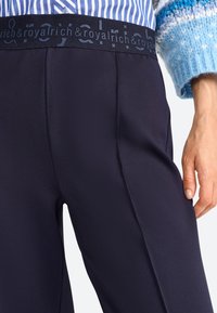 Pantaloni a vita alta blu navy con tessuto testurizzato e una cintura con logo scuro. Dettagli delle cuciture visibili e vestibilità aderente nella zona delle cosce.