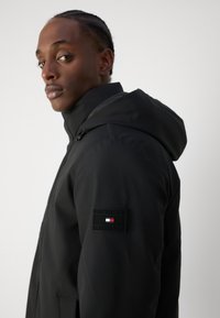 Schwarze Kapuzenjacke aus strukturiertem Stoff, mit einem auffälligen Logo-Patch im rot-weiß-blauen Design auf dem linken Ärmel.