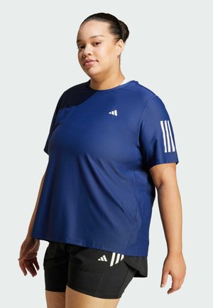 Osoba w niebieskiej sportowej koszulce z białymi paskami na rękawie i czarnych szortach z białymi logo, stojąca na jednolitym tle.