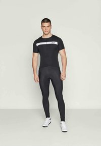 Modello maschile che indossa una maglia da ciclismo attillata bianca e nera e leggings da ciclismo neri con scarpe da ginnastica bianche, in piedi di fronte a uno sfondo semplice.