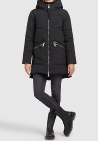 Manteau rembourré noir avec capuche, fermeture éclair et deux poches zippées. Présente une longueur de dos plus longue et un design épuré. Assorti avec un jean foncé et des bottes noires.