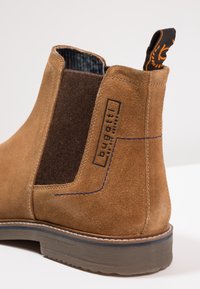 Hnědé semišové kotníkové boty s tmavě hnědými plstěnými bočními panely, modrými švy a dekorativním poutkem s oranžovým vzorem. Plná gumová podrážka.