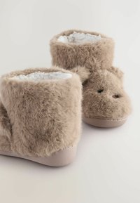 Beige, flauschige Hausschuhe mit einer weichen Textur, runder Form, umschlagbarem Design und kleinen dunklen Akzenten für die Augen auf der Vorderseite.