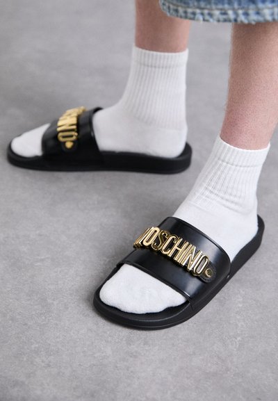 Person iført hvide sokker med sorte slidesandaler, der har guld "Moschino" branding, på grå gulv.